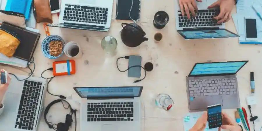 An overhead shot of a busy workspace with multiple laptops, smartphones, and office supplies scattered around a wooden table.